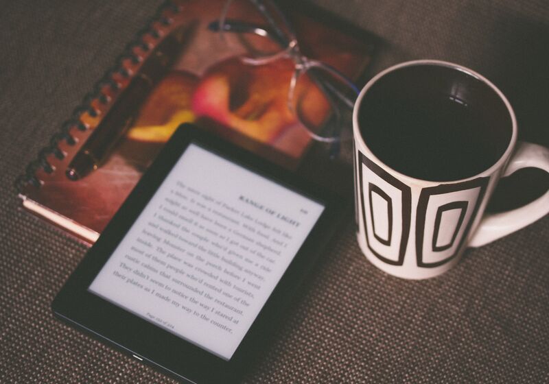An e-reader placed next to a mug, reading glasses and notebook and pen